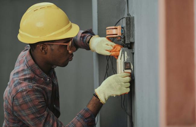 A man in a hardhat and gloves installing equipment on a wall.